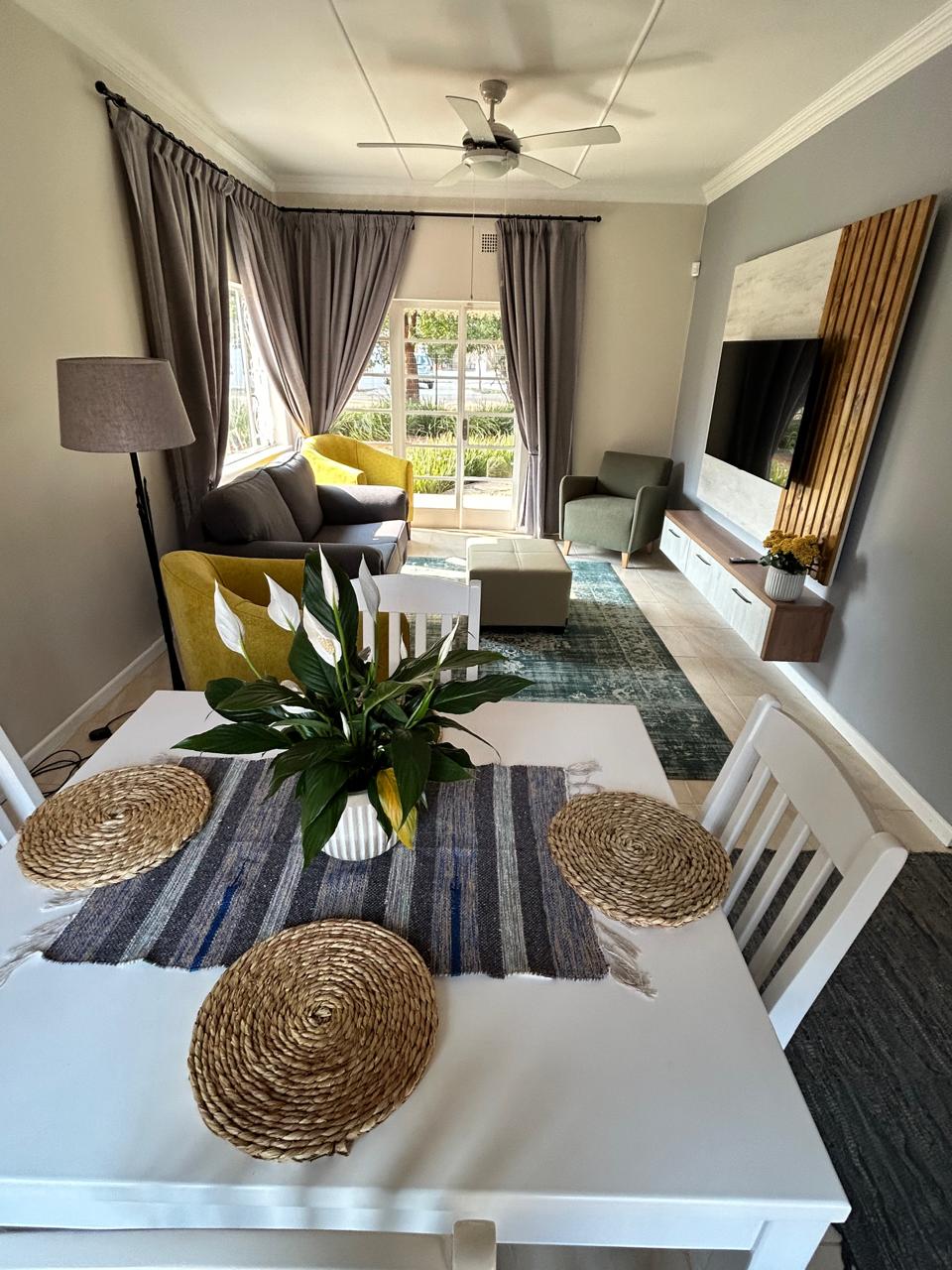 A modern living area features a comfortable yellow couch and a grey armchair, framed by large windows with flowing curtains. A tasteful dining table with woven placemats sits at the forefront, complemented by a decorative plant. A flat-screen TV is mounted on the wall.