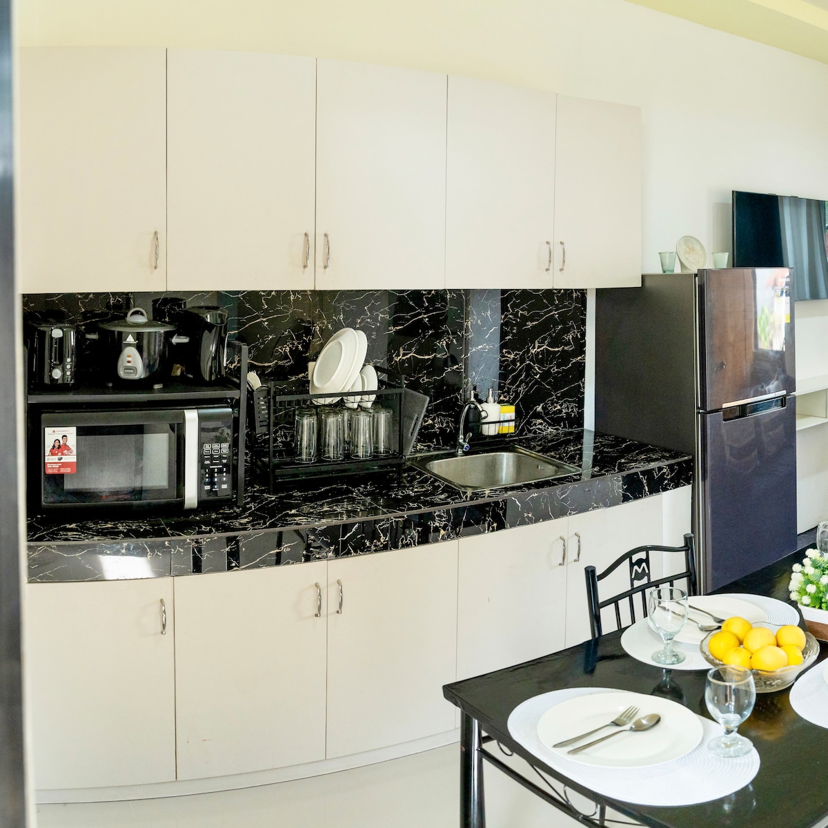 A modern kitchen features black marble countertops and light cabinetry, showcasing a microwave, coffee maker, and dishware. A small sink is situated near the appliances, while a black dining table is set with plates and fresh fruit, creating a functional and inviting dining space.