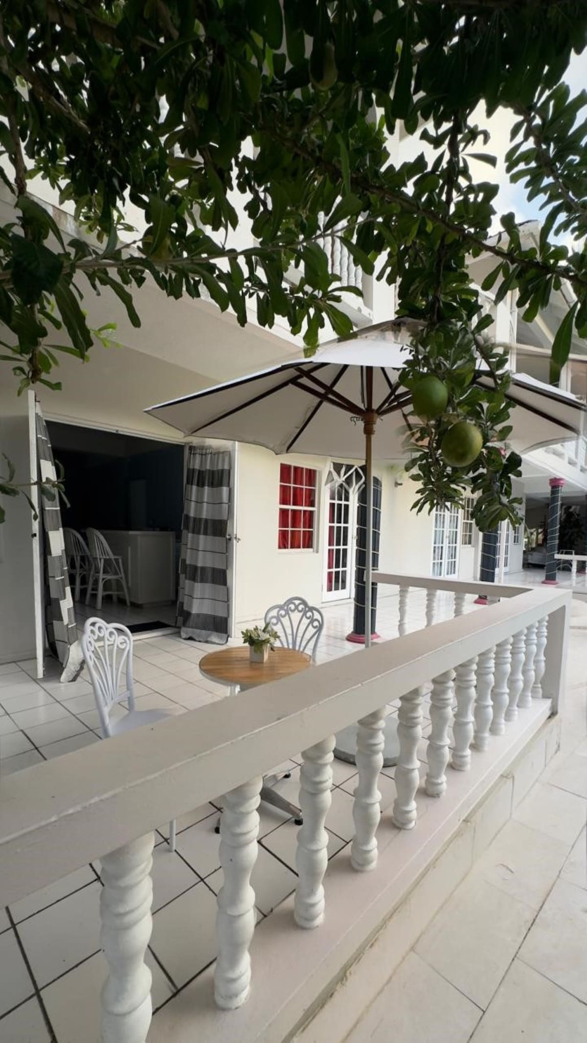 A small outdoor seating area is depicted, featuring a round wooden table with two white chairs. An umbrella provides shade, while greenery frames the scene. The building's features include white columns and multiple doors with decorative windows offering glimpses of the interior.