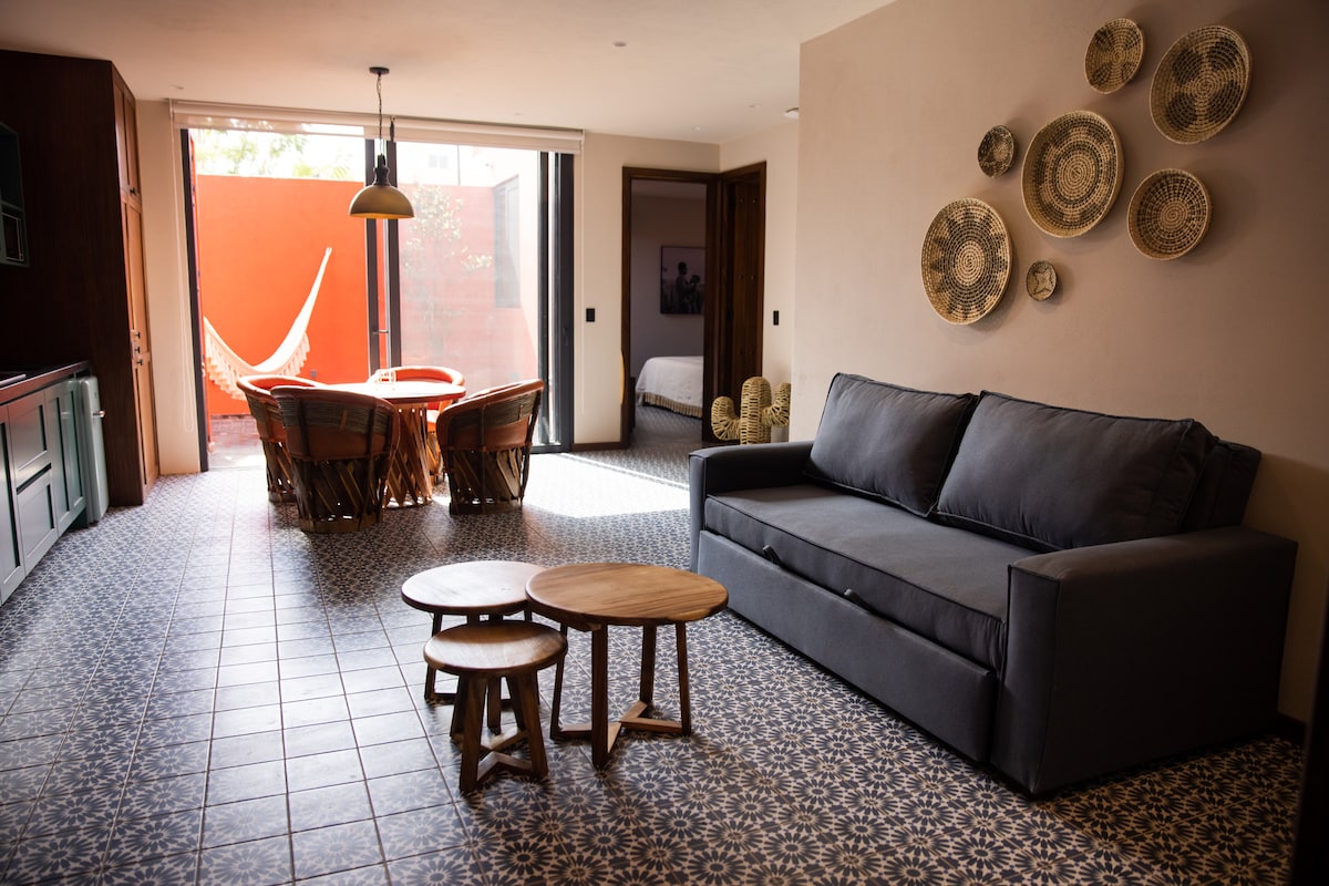A comfortable living area features a dark gray sofa facing two round wooden tables. Behind, a dining area with woven chairs is visible beside large glass doors that lead to an outdoor space. Decorative wall baskets complement the earthy tones of the room.