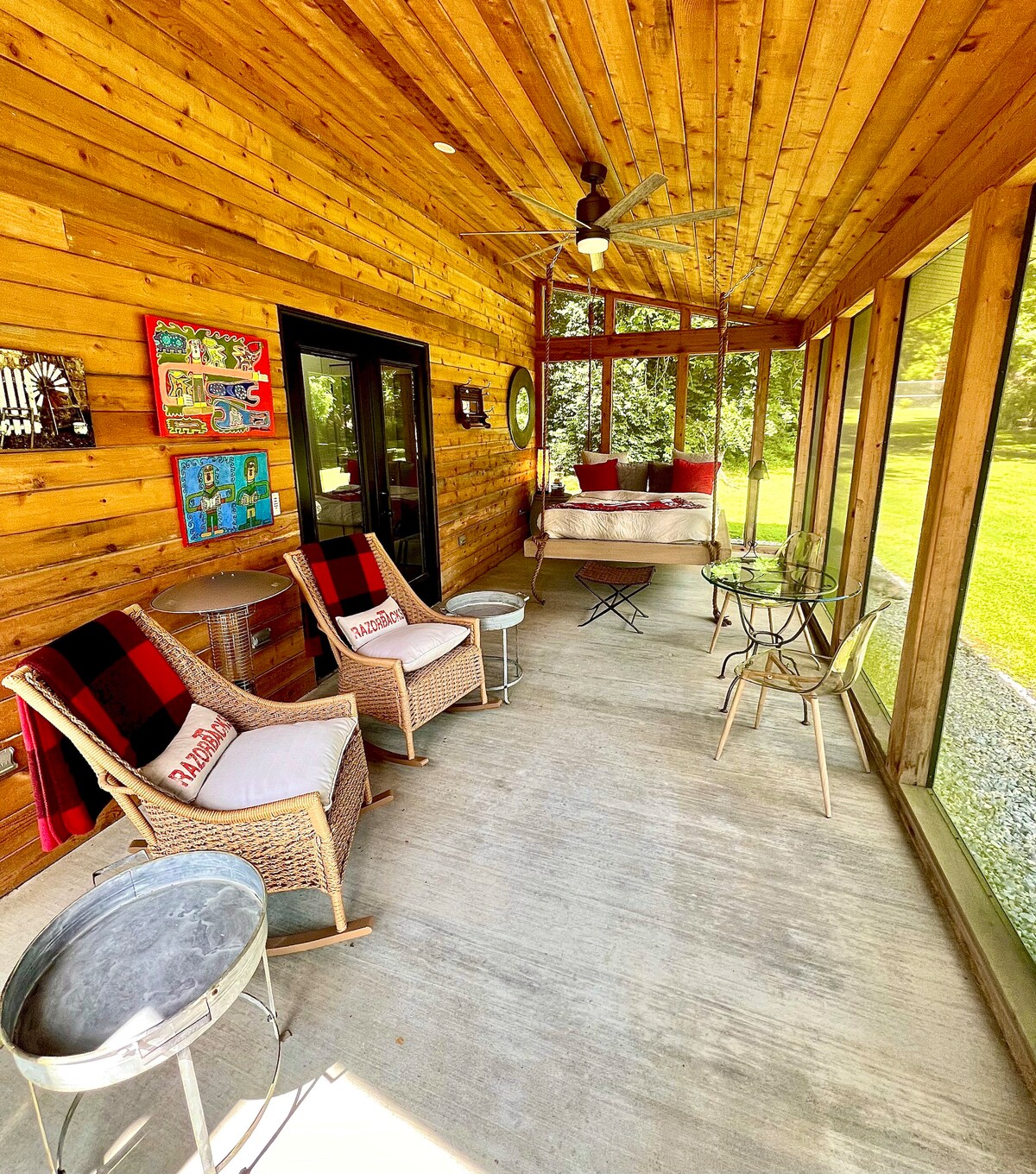 A screened-in porch is showcased, featuring a spacious swing bed and two wicker chairs with red and black plaid cushions. A small glass table and metal side table are present, set against a backdrop of wooden walls and views of the green outdoors.