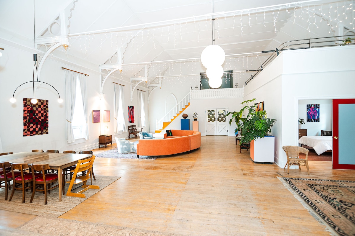 The spacious living area features high ceilings and large windows, allowing natural light to fill the room. A round dining table is surrounded by wooden chairs, while a colorful curved sofa invites relaxation. Artwork adorns the walls, enhancing the artistic atmosphere of the space.