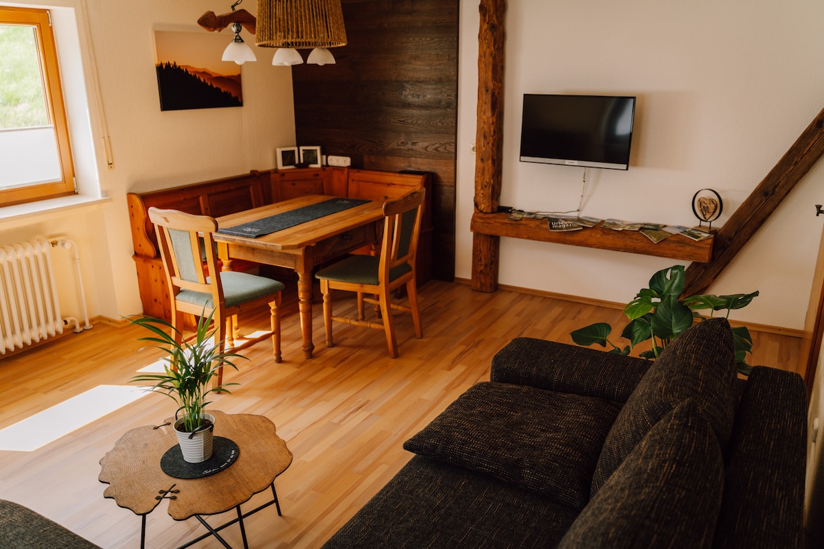 A living and dining area is featured, showcasing light wooden flooring and warm wall textures. A dining table surrounded by four chairs is positioned beside a corner bench. A television is mounted on the wall, complemented by a small coffee table and potted plant near the seating area.