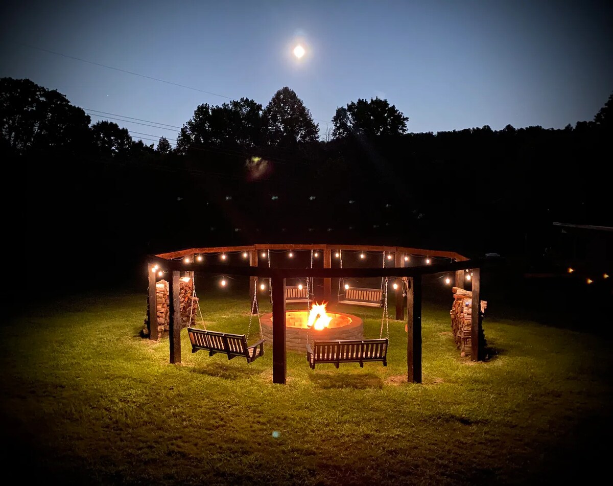 A circular gathering area is illuminated by string lights, surrounding a central fire pit with a vibrant flame. Two wooden swings are positioned beside the fire, and the soft glow of the moon is visible in the night sky above the tree line.