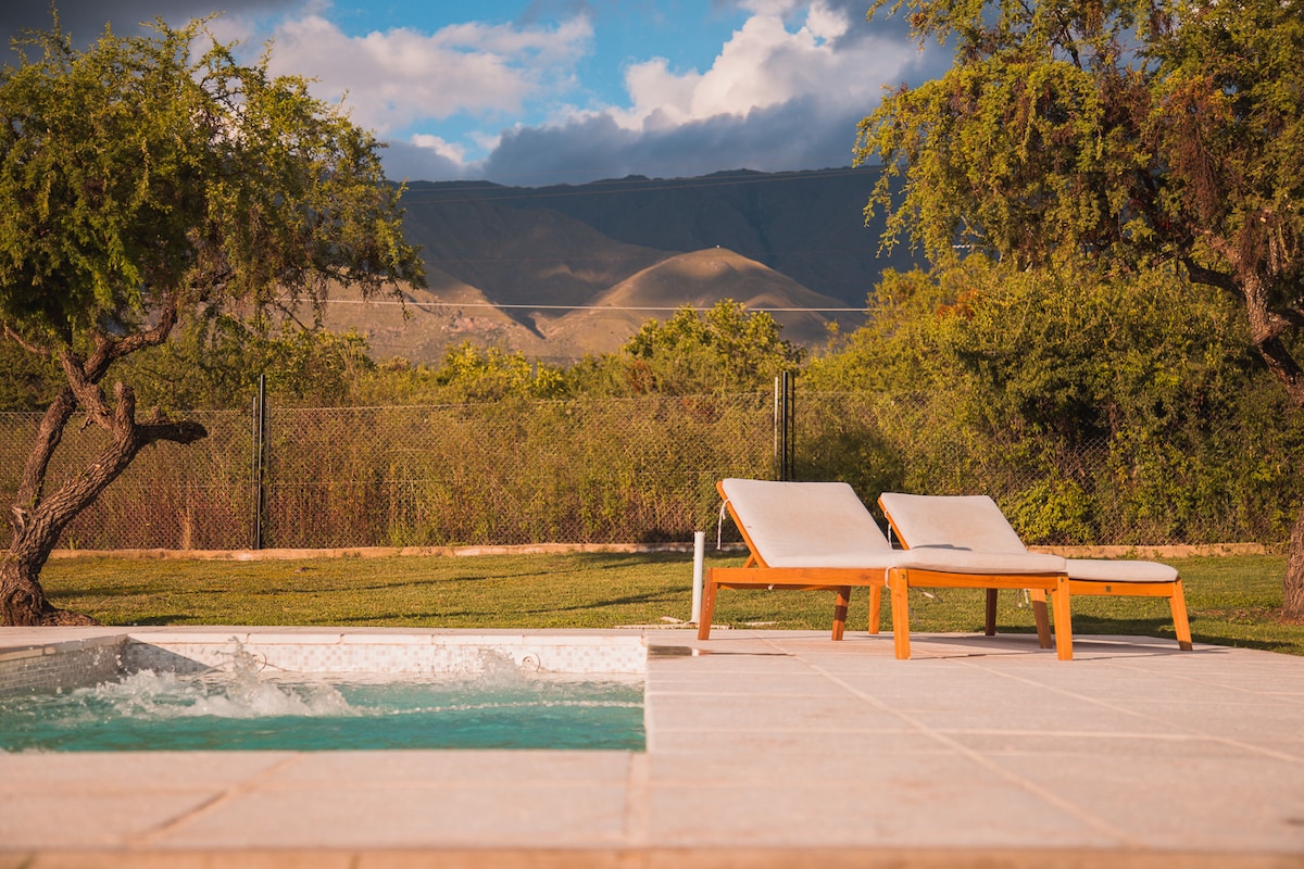 Lounge chairs are positioned beside a private pool, with a soothing whirlpool feature. The backdrop displays rolling mountains and lush greenery, creating a serene outdoor setting. Soft sunlight bathes the area, enhancing the tranquil atmosphere.