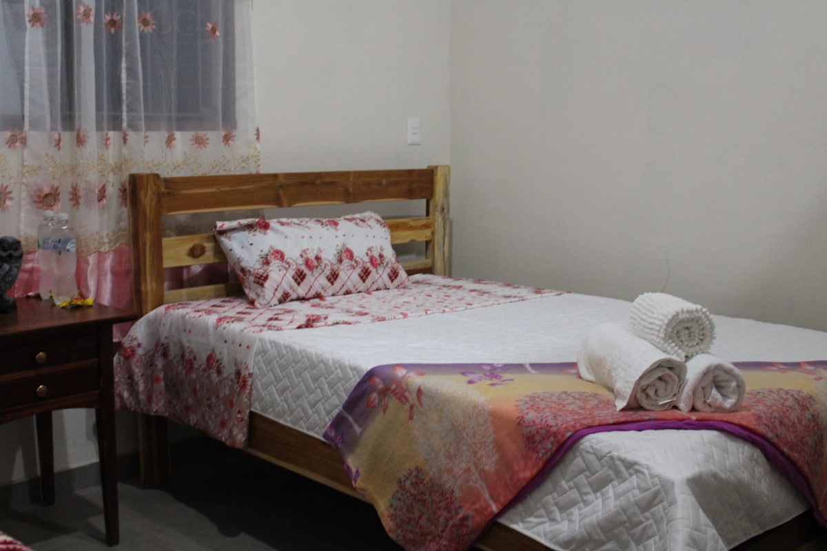 A cozy sleeping area features a wooden bed adorned with floral-patterned bedding and decorative cushions. Fresh towels are neatly arranged at the foot of the bed. A bedside table stands next to the bed, while a window with sheer curtains allows natural light to enter the room.