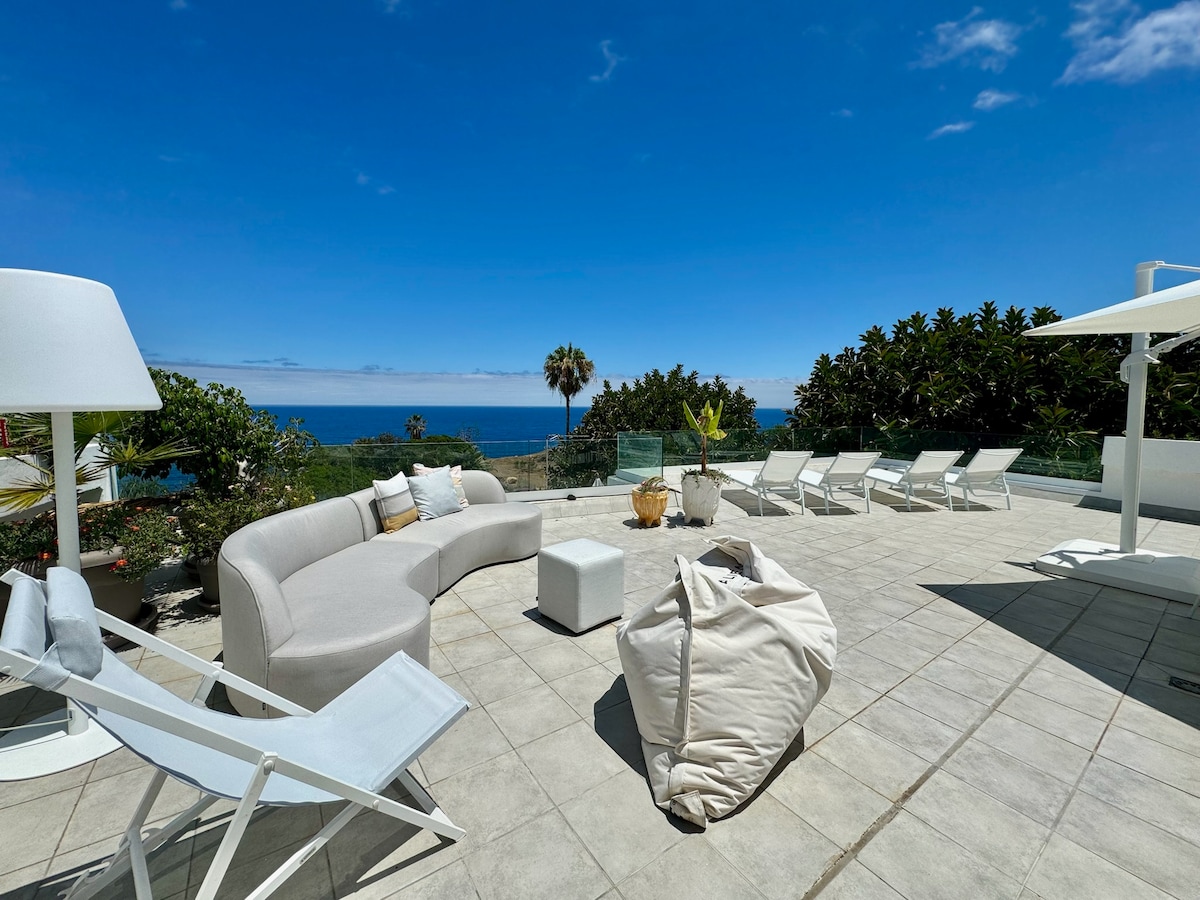 The spacious patio features outdoor seating with a curved sofa and a large white bean bag. Sun loungers are arranged around the area, offering views of the ocean and gardens. A shaded dining area is provided with a stylish umbrella, creating a relaxing environment.