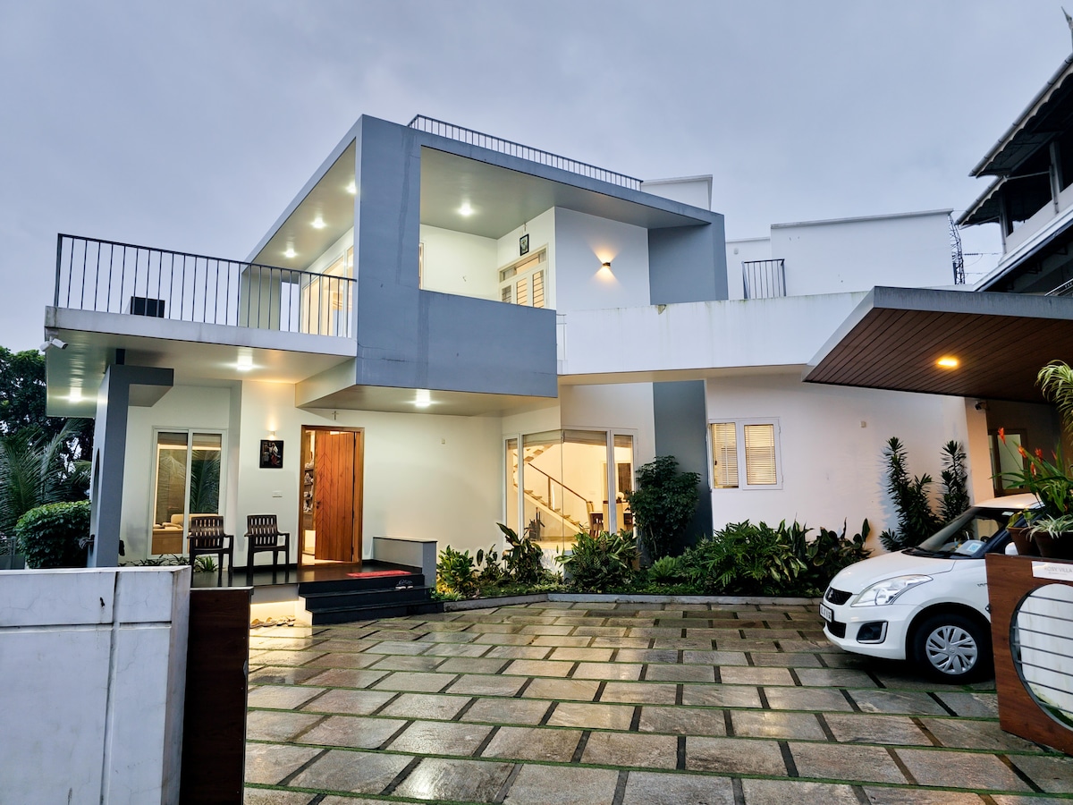 The modern villa exterior features clean lines and multiple balconies, surrounded by well-maintained landscaping. A paved driveway leads to a covered entrance, with outdoor seating visible beside the main entrance. The light fixtures illuminate the home, enhancing its evening charm.