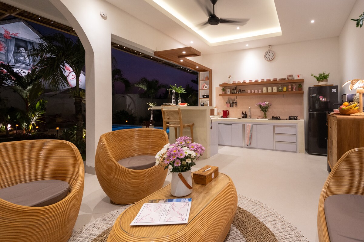 A serene common area features a stylish kitchen and seating arrangement. Curved wicker chairs surround a circular table, while a view of the garden and pool is visible through the open windows. Soft lighting enhances the welcoming environment, creating a cozy space for relaxation.