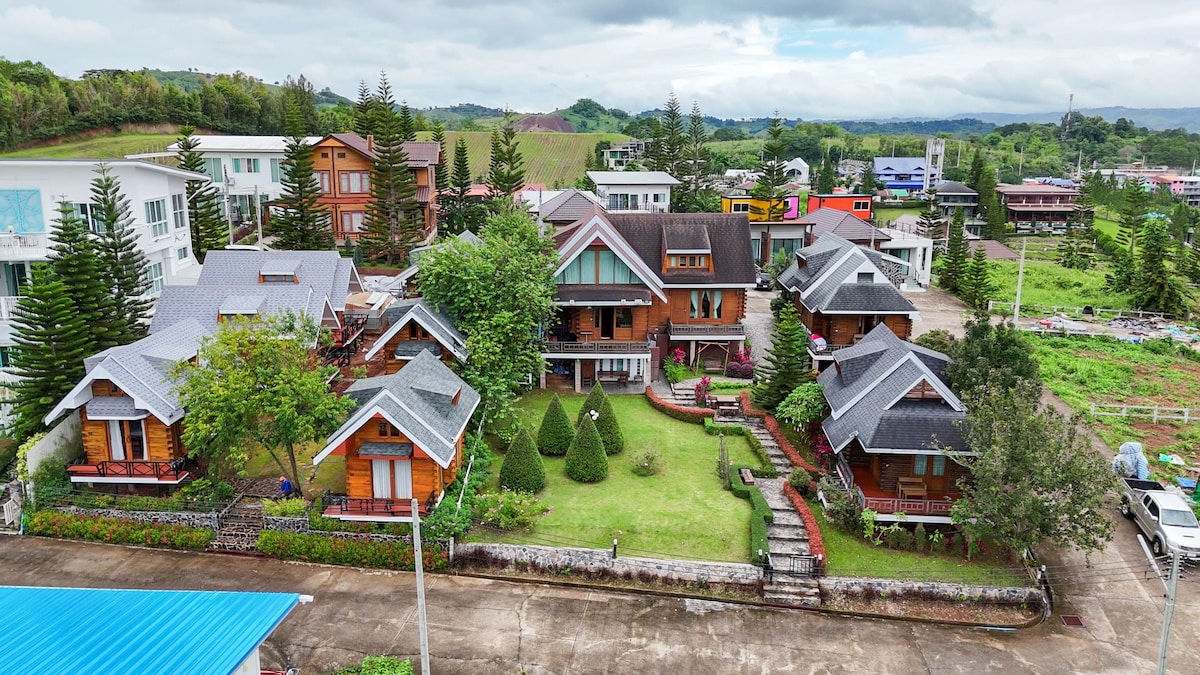 Aerial view of a wooden resort surrounded by lush greenery. Multiple cabins with sloped roofs and balconies are situated around a manicured lawn, featuring neatly trimmed bushes and pathways. The backdrop consists of gentle hills and a clear sky, enhancing the natural setting.