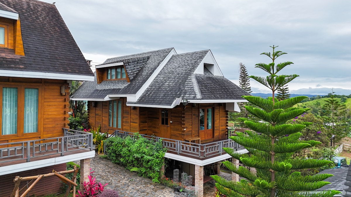A wooden resort is shown nestled among natural surroundings, featuring a sloped roof and multiple levels. Lush greenery and colorful plants enhance the exterior, while a stone pathway leads to the entrance. Views of distant hills are visible beyond the resort.