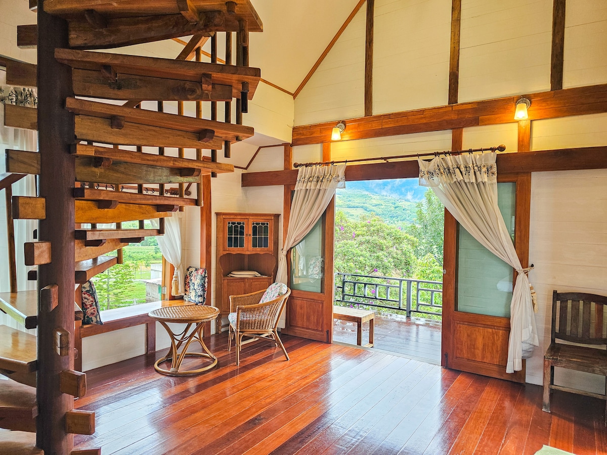 An open living space features wooden flooring and large glass doors that reveal scenic mountain views. A spiral staircase made of wood leads to an upper level. Multiple seating options, including a cozy chair and a wicker table, offer relaxation areas, framed by soft curtains.