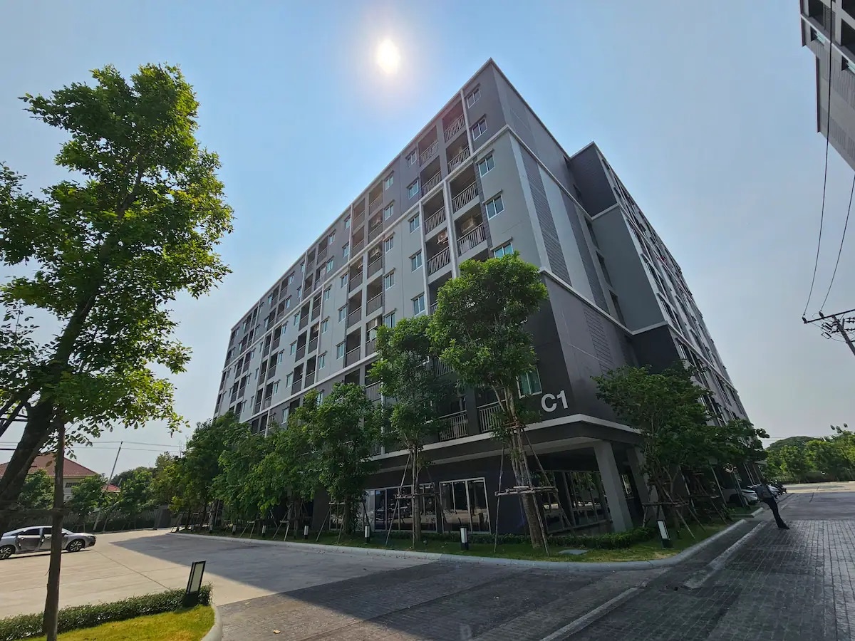 A modern four-story condominium complex is viewed from the ground. The facade is framed by greenery, with sunlight casting a warm glow. The surrounding driveway features smooth paving and landscaped areas, providing an inviting entrance to the building.
