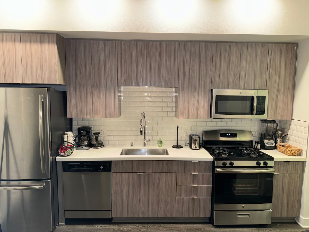 The kitchen features modern cabinetry with a light wood finish, complemented by a sleek countertop. Stainless steel appliances, including a gas stove, microwave, and dishwasher, are present. A coffee maker and a toaster are also visible, along with a woven basket and various kitchen utensils.