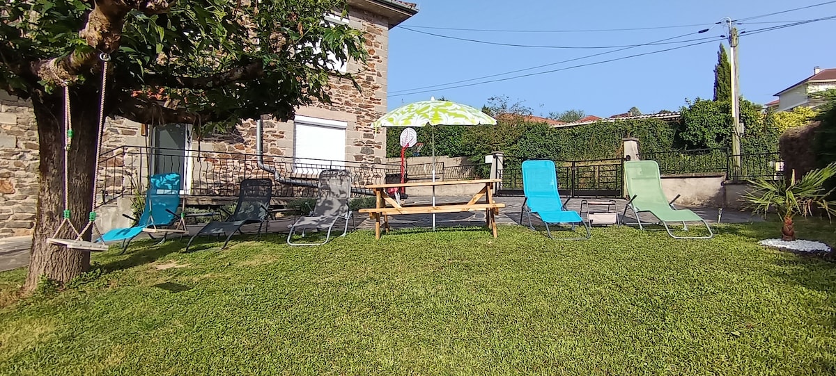 A grassy outdoor area is presented, featuring multiple lounge chairs in various shades and a large picnic table. An umbrella offers shade, while a backdrop of a stone building and greenery enhances the space. The area appears ideal for relaxation and social gatherings.