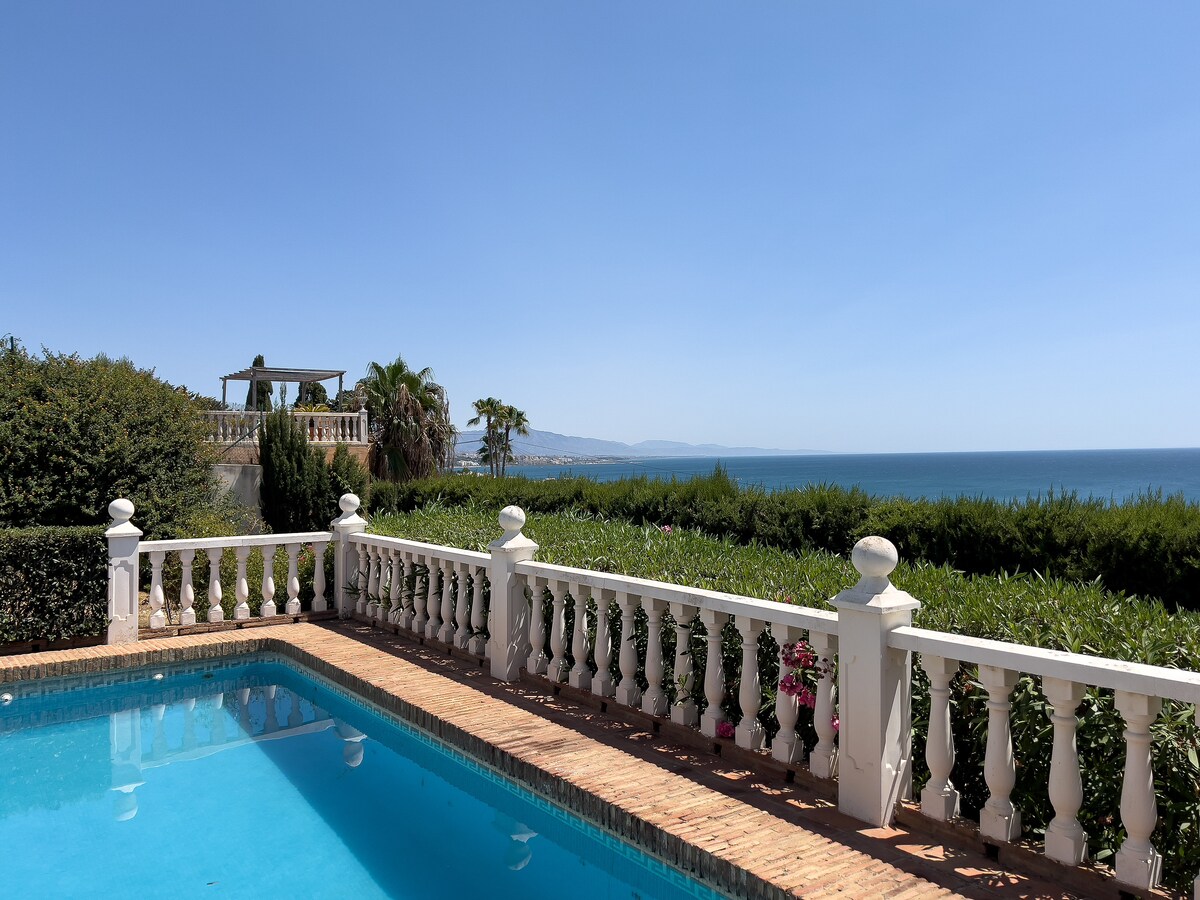 A serene outdoor area is showcased, featuring a clear blue pool surrounded by decorative white railings. Lush greenery and palm trees extend towards the horizon, where the ocean meets the sky. A pergola is visible in the background, inviting relaxation.