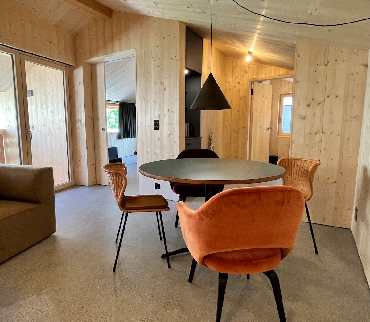 A dining area is featured with a round table surrounded by four chairs in warm colors. Natural wood paneling creates a cozy ambiance. Large glass doors allow for natural light and a view of the surrounding area, enhancing the inviting space.
