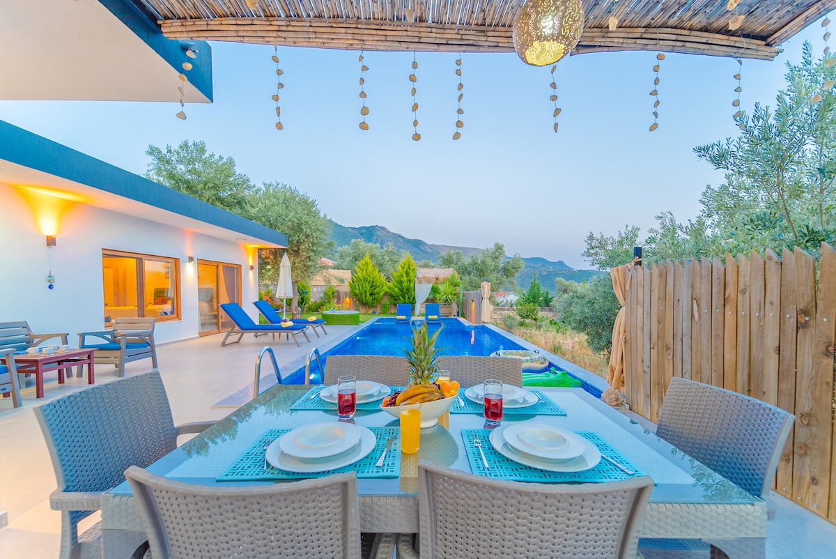 An outdoor dining area is set with a table and chairs, featuring dishes and drinks. In the background, a swimming pool is visible, surrounded by greenery and distant mountains. Sun loungers are arranged nearby, creating an inviting space for relaxation.