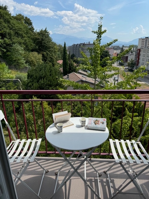A small balcony features a round white table accompanied by two foldable chairs. A book and two cups are placed on the table. Lush greenery lines the view, with distant mountains visible under a blue sky dotted with clouds.