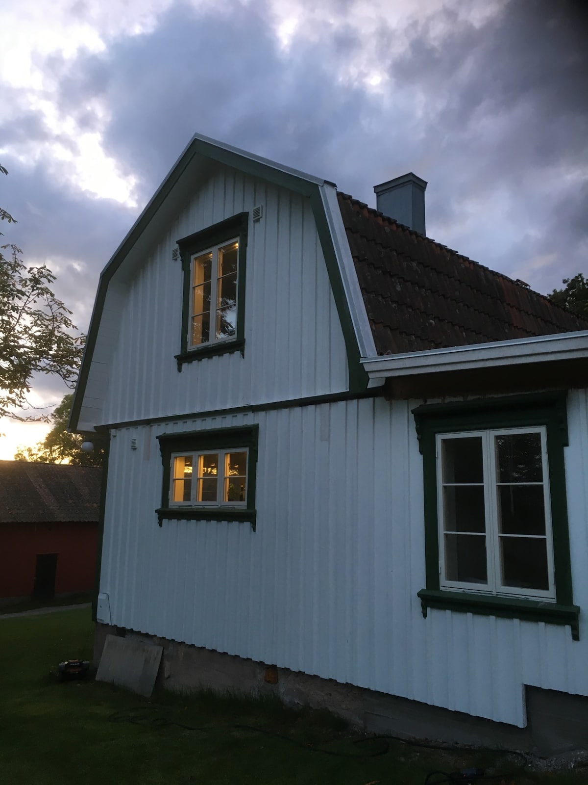 The exterior of a traditional house is shown, featuring white wooden siding and green trim. Two windows reflect warm light from within, and the roof is covered with dark tiles. A cloudy sky is visible overhead, adding to the serene ambiance of the setting.