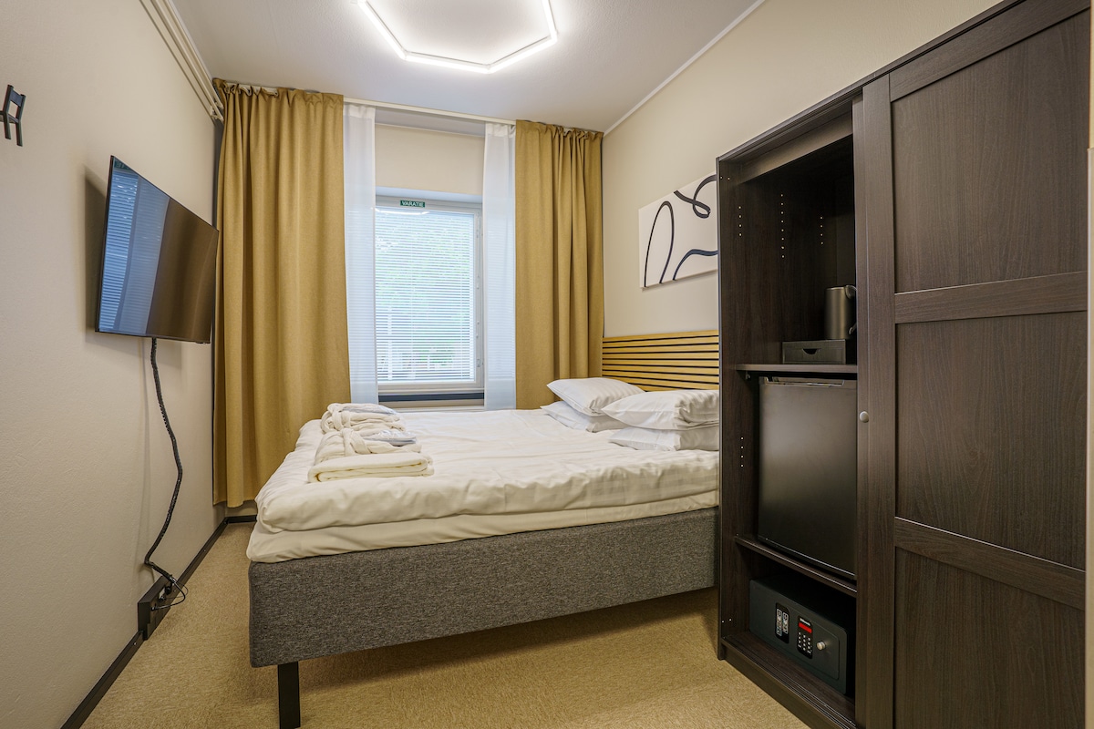 The room features a spacious bed with soft bedding, a wall-mounted television, and a dark wood cabinet. Natural light enters through the window, which is dressed with sheer and solid curtains. Amenities include a kettle and a safe, enhancing convenience.