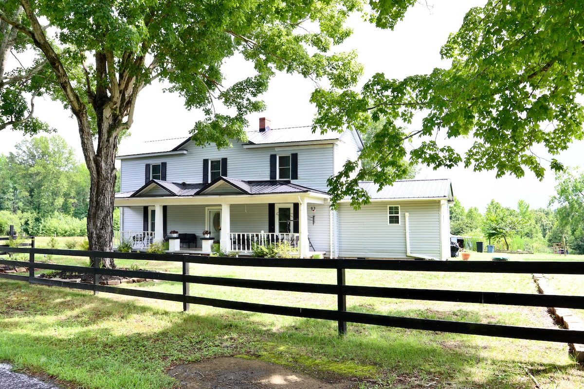 The exterior of a refurbished farmhouse is showcased, featuring a light-colored facade and a charming front porch. A large tree provides shade, while a black fence borders the property. The surrounding landscape reveals lush greenery and open fields.