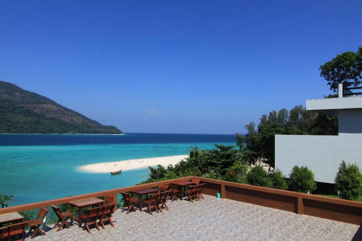 A spacious terrace offers a view of a sandy beach bordered by clear turquoise waters. A mountain rises in the background, and several wooden chairs are arranged for outdoor seating, surrounded by greenery.