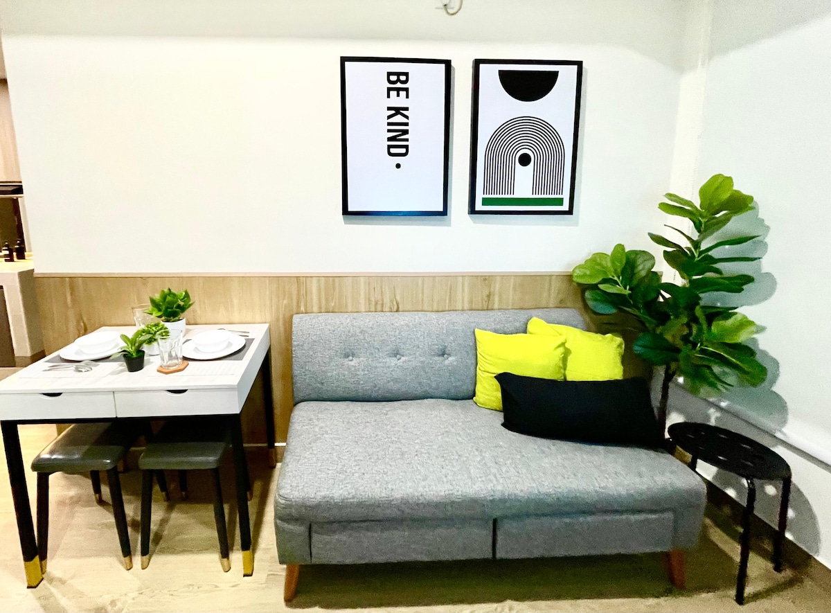 A cozy corner features a light gray sofa adorned with vibrant green and black pillows. A small dining table with two chairs sits nearby, dressed with plates and a small plant. Wall art showcasing simple, inspirational phrases complements the warm design.