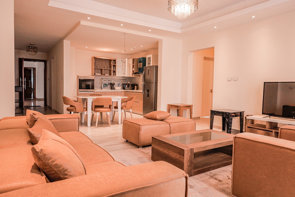 A spacious living area is furnished with comfortable brown seating and a coffee table. The open-concept layout includes a dining area with a modern table and chairs, as well as a kitchen visible in the background with sleek cabinetry and appliances.