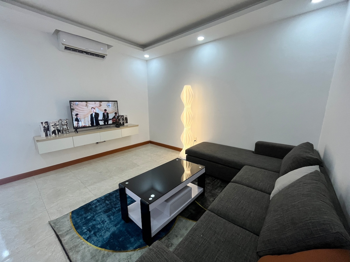 A comfortable living area is furnished with a dark sectional sofa, centered around a sleek coffee table. A wall-mounted television is positioned above a minimalist entertainment unit. Soft lighting and a contemporary floor lamp create a cozy ambiance.