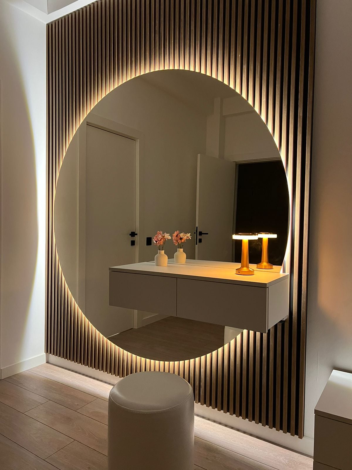 A large, oval mirror is framed by vertical wooden slats, creating a subtle background. A minimalist console table sits below, adorned with two vases holding fresh flowers and a modern table lamp with a warm glow. Neutral tones are complemented by soft lighting.