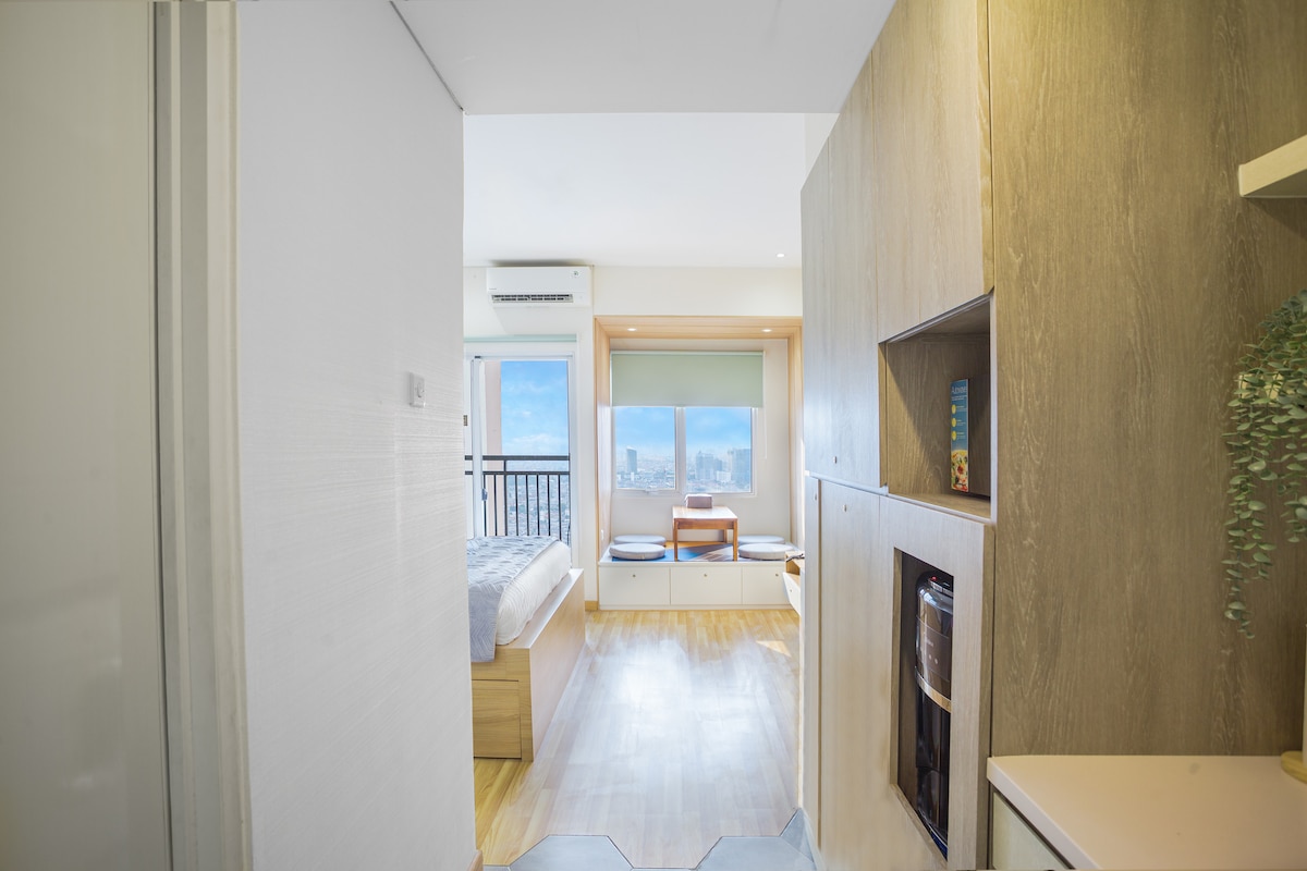 A bright hallway leads into a studio apartment with wooden flooring. The spacious living area features a large window offering a cityscape view. Minimalist furniture, including a bed with soft linens and seating by the window, creates a functional and inviting atmosphere.