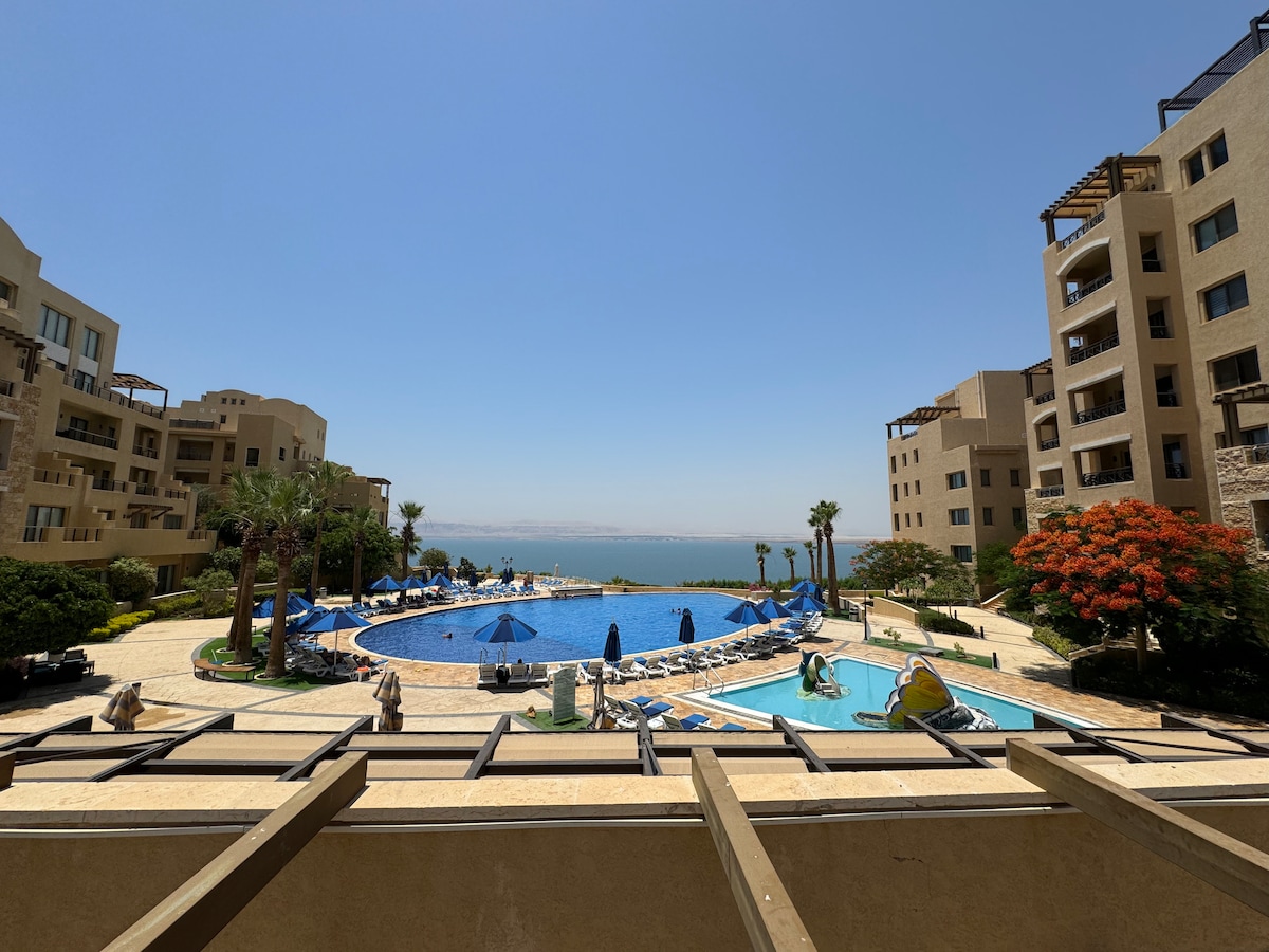 The view features a large blue pool surrounded by lounge chairs and umbrellas. Palm trees line the area, leading down to the Dead Sea, which glimmers under the clear sky. The complex's buildings are visible in the background, enhancing the serene landscape.