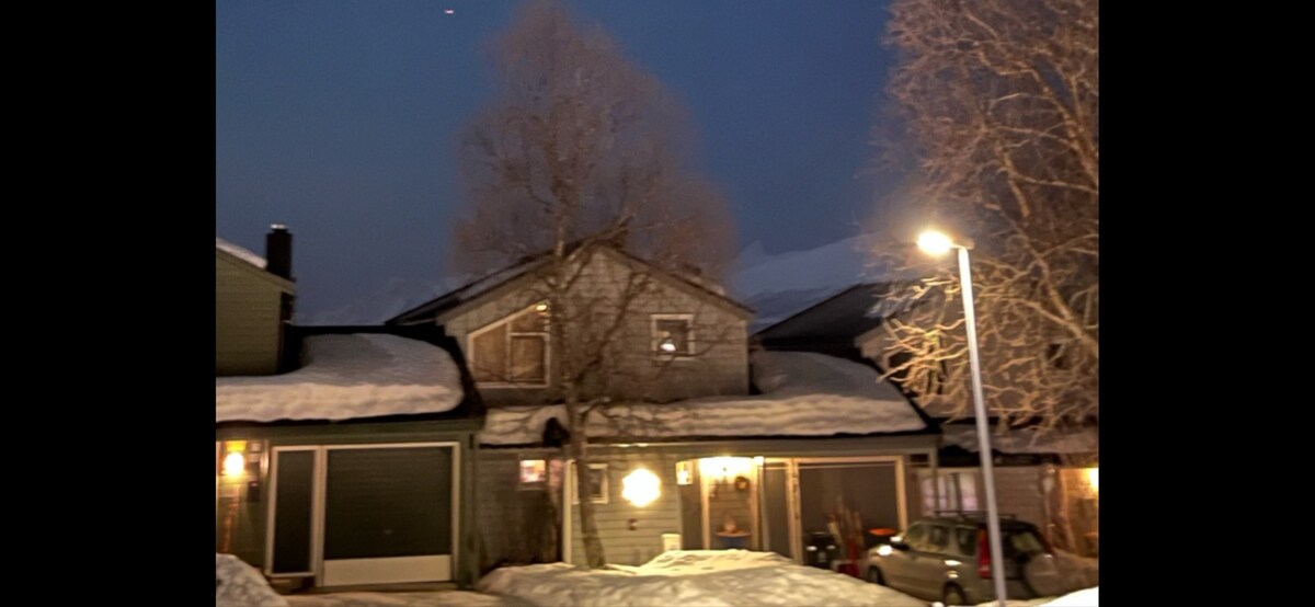 The exterior of a two-story house is shown during twilight, with snow blanketing the roof. Warm light spills from a window, and trees bare of leaves are visible in the foreground. A parked car and a sidewalk lead up to the entrance.