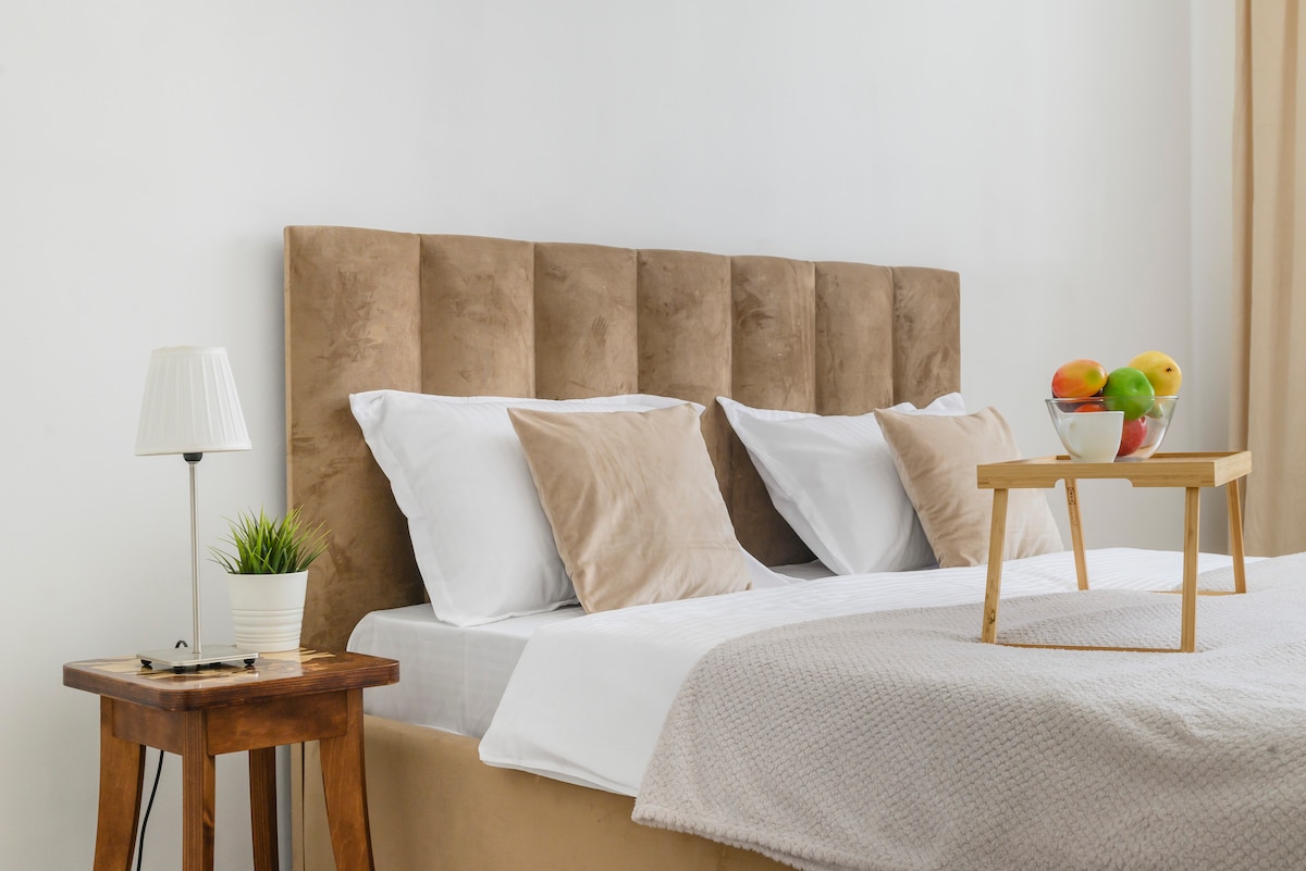 A spacious bed is adorned with soft pillows and a neutral-colored blanket. A small wooden table holds a fruit bowl, and a bedside lamp provides gentle illumination. A potted plant adds a touch of greenery to the inviting space.