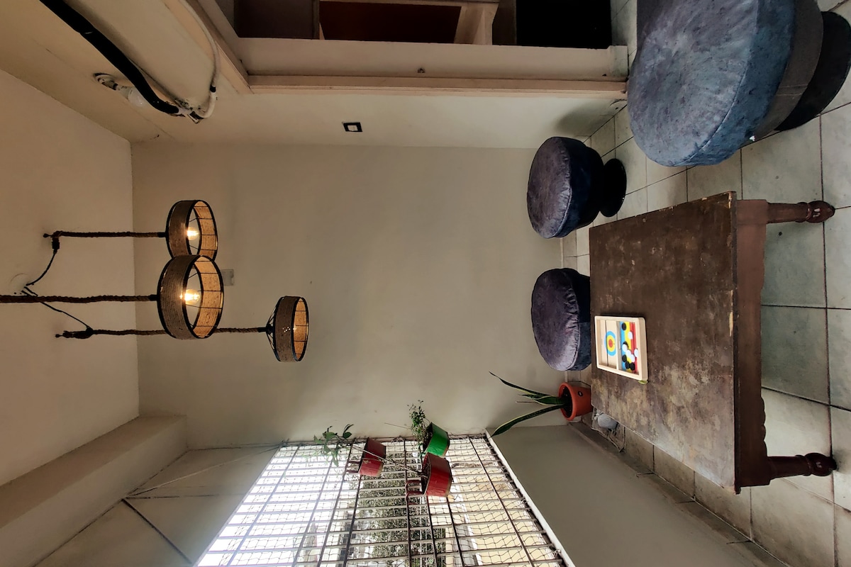 A cozy seating area is arranged with a wooden coffee table and circular poufs covered in soft fabric. Three pendant lights hang from the ceiling, casting a warm glow. Light filters through a large window partially obscured by greenery from potted plants placed nearby.