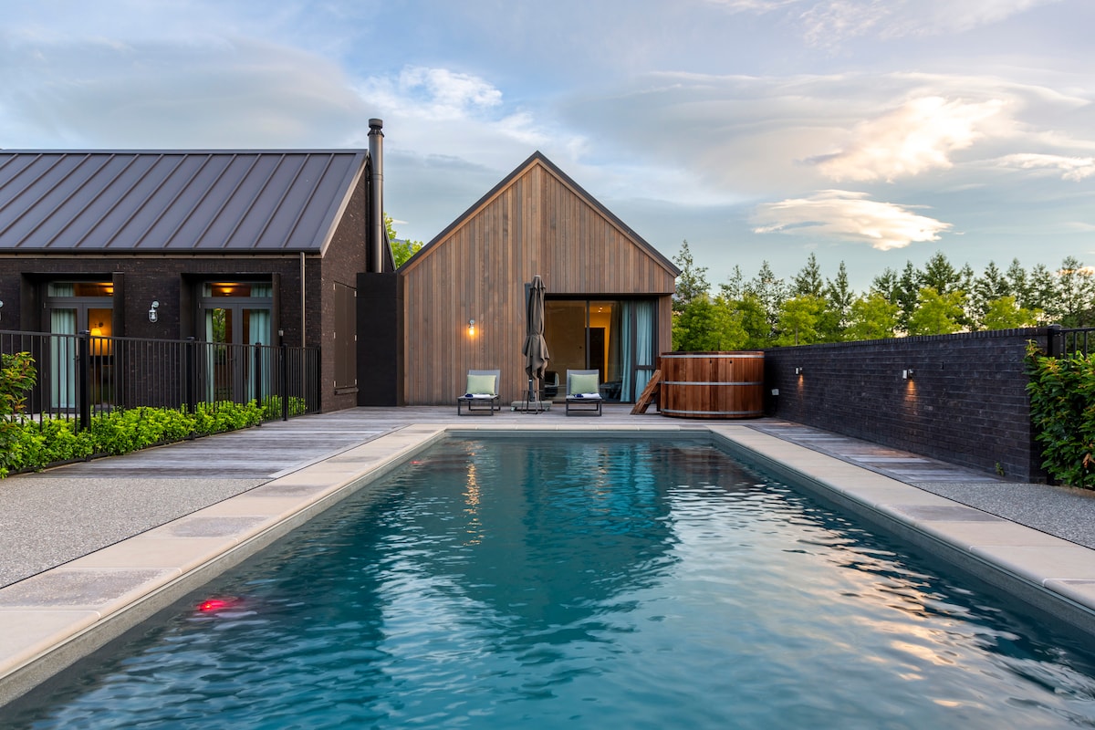 A modern villa features a private pool surrounded by a tiled deck. Lounge chairs are positioned near the water, and the structure showcases a mix of dark and natural wood exteriors. Green landscaping frames the area, providing a serene outdoor retreat.