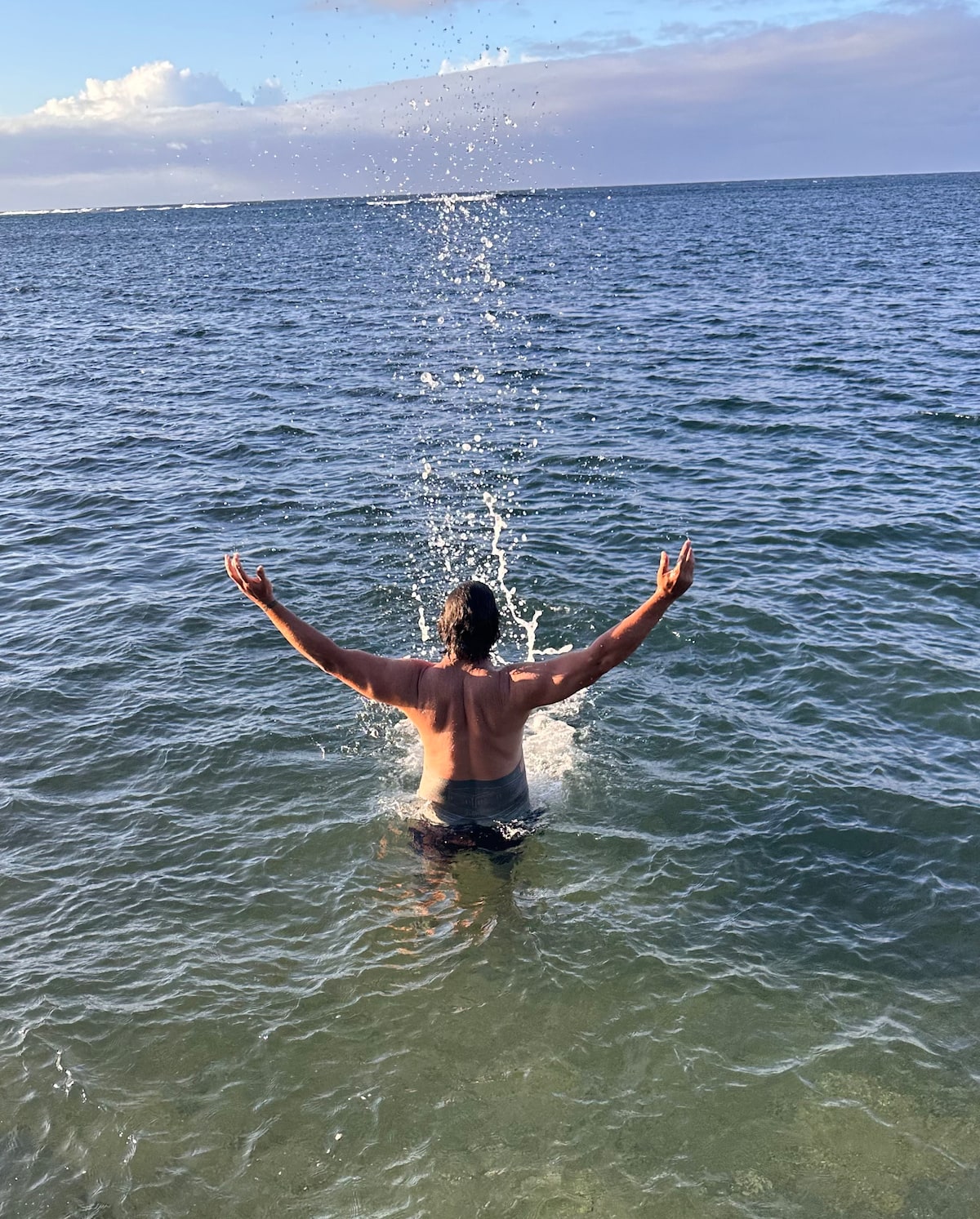 A person is standing in shallow water, with arms raised joyfully. Water splashes around them, reflecting the sunlight. The background shows a calm sea merging with a soft horizon under a partly cloudy sky.