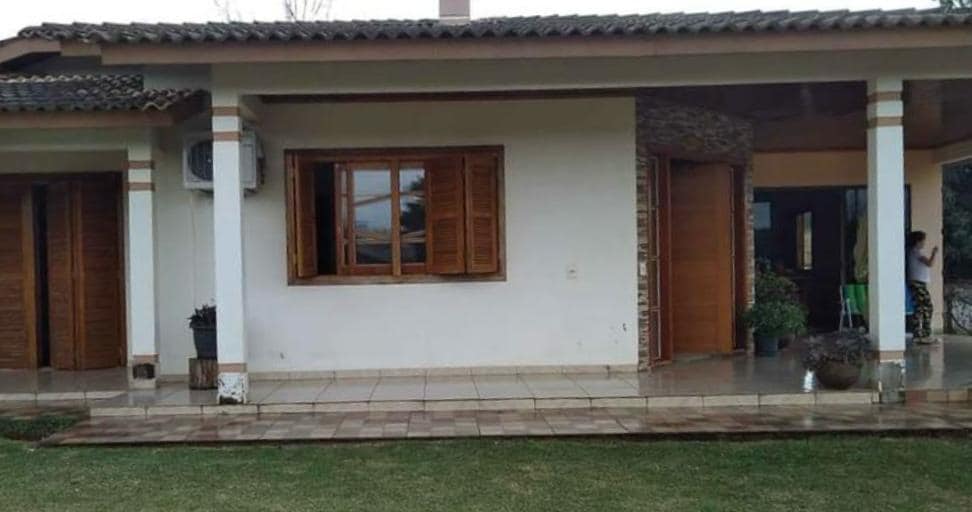 The exterior of the house is depicted, featuring a covered porch with wooden shutters on the window. A tiled pathway leads to a well-maintained garden area, framed by green grass and potted plants. The entrance showcases a door made of natural wood.