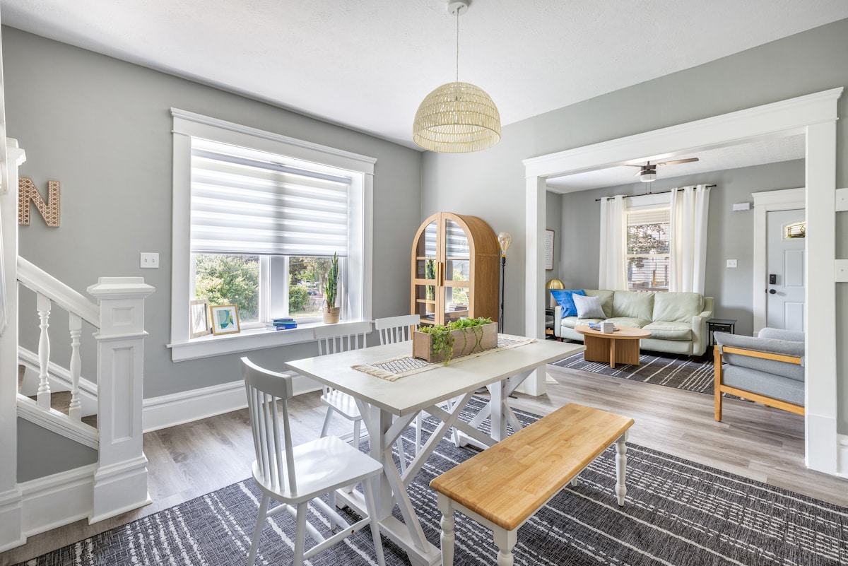 A dining area features a white table with a wooden bench and chairs, complemented by a large window allowing natural light. An arched wooden room divider leads to a cozy living space visible in the background, contributing to an open and welcoming environment.
