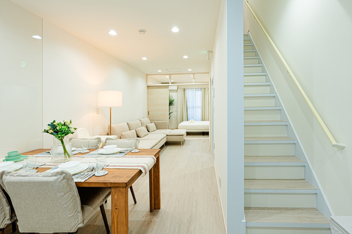 An open living area is visible, featuring a light-toned wooden dining table set for six, and a cozy seating arrangement with a large sectional sofa. A staircase leads to the upper level, while natural light enters through a window, enhancing the bright and airy space.