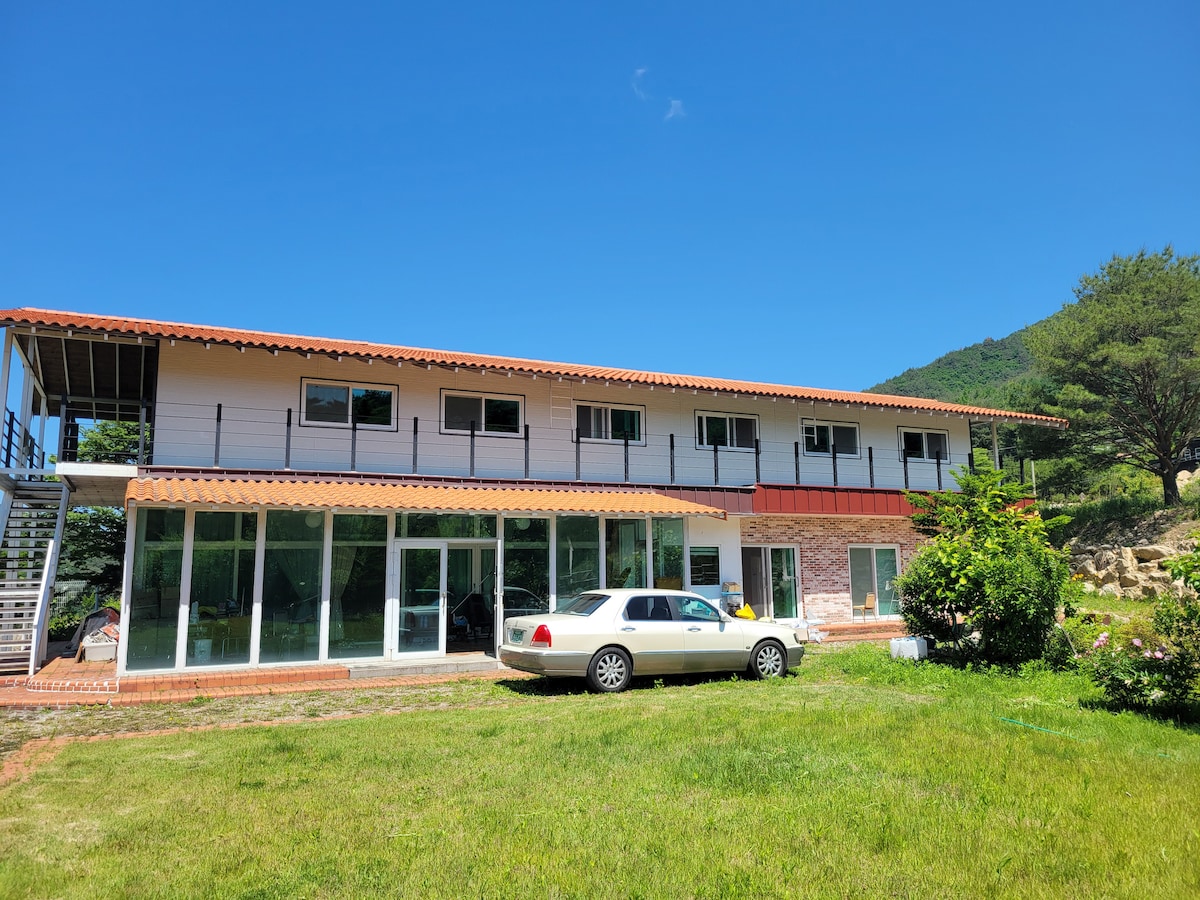 A spacious two-story building is set against a clear blue sky, featuring large glass windows that allow natural light to flood the interior. Lush green grass surrounds the property, with a car parked in front, and a mountain is visible in the background.