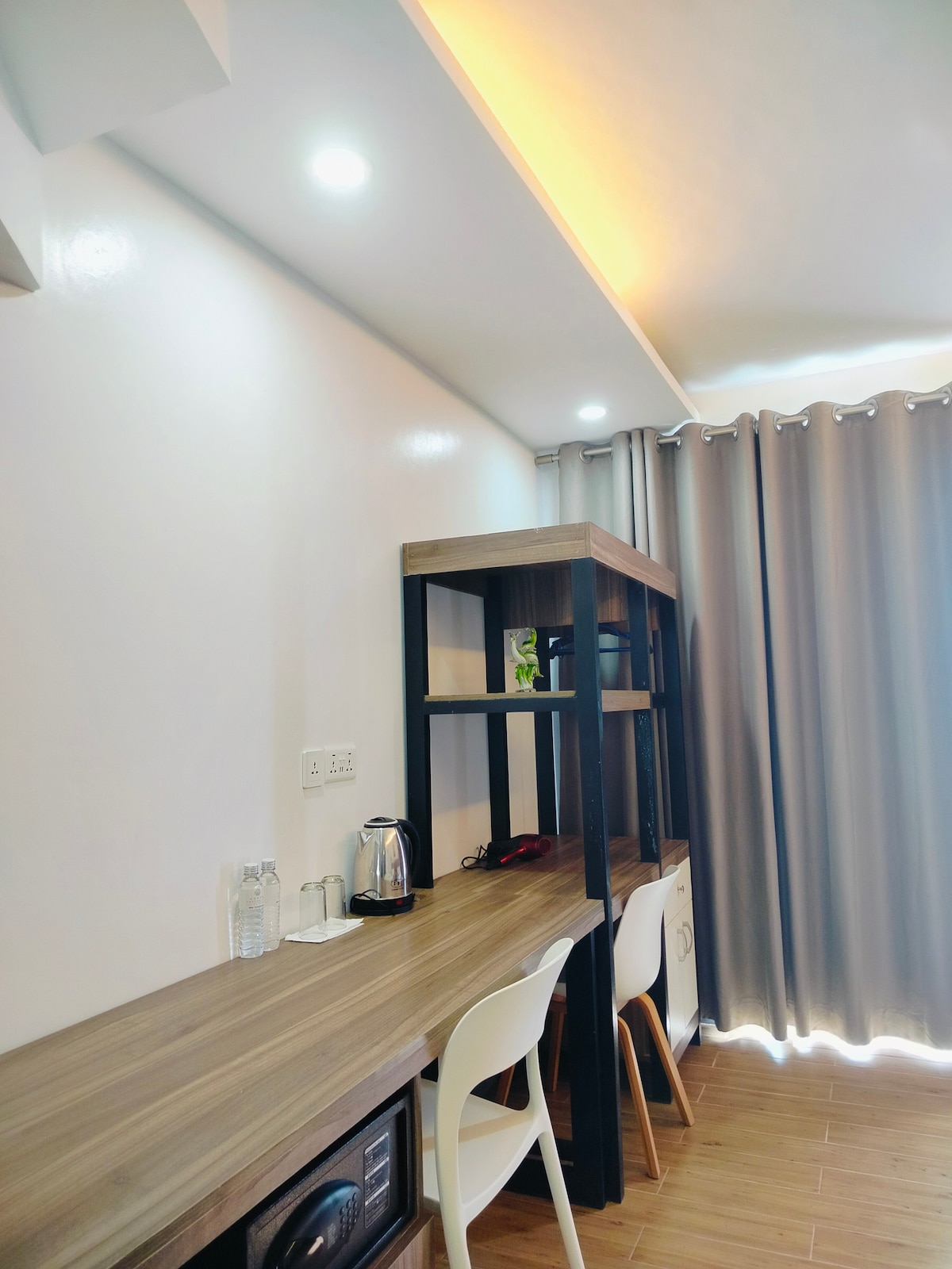 A modern workspace is presented featuring a wooden desk against a light-colored wall. A kettle and two bottles of water sit on the desk, while a small plant adds a touch of greenery. The area is illuminated by recessed ceiling lights and natural light filtering through gray curtains.