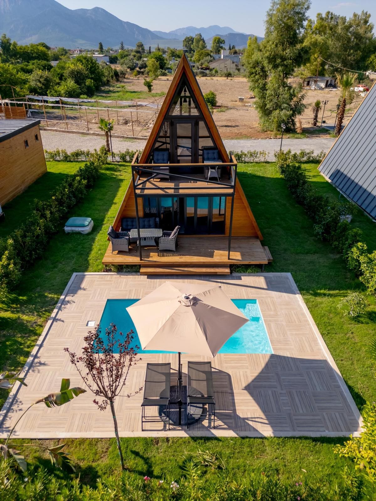 An A-frame villa is surrounded by greenery, featuring a private pool with lounge chairs and a large umbrella on a stone patio. The outdoor area offers a view of the mountains in the background, complemented by a well-maintained lawn.