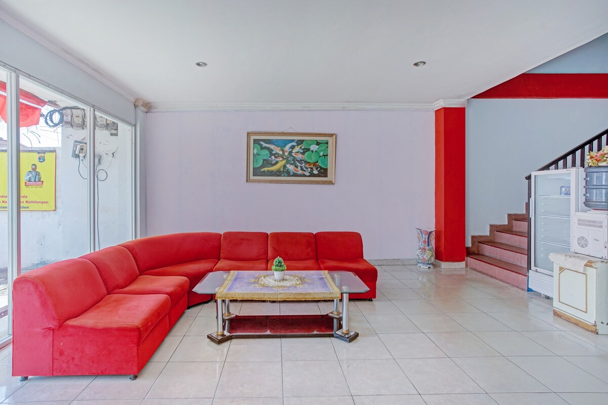 A vibrant seating area is highlighted by a bold red sectional couch, arranged around a glass-topped coffee table. The room features a large window allowing natural light, with decorative artwork displayed on the wall. A staircase leads to an upper level, complementing the open space.