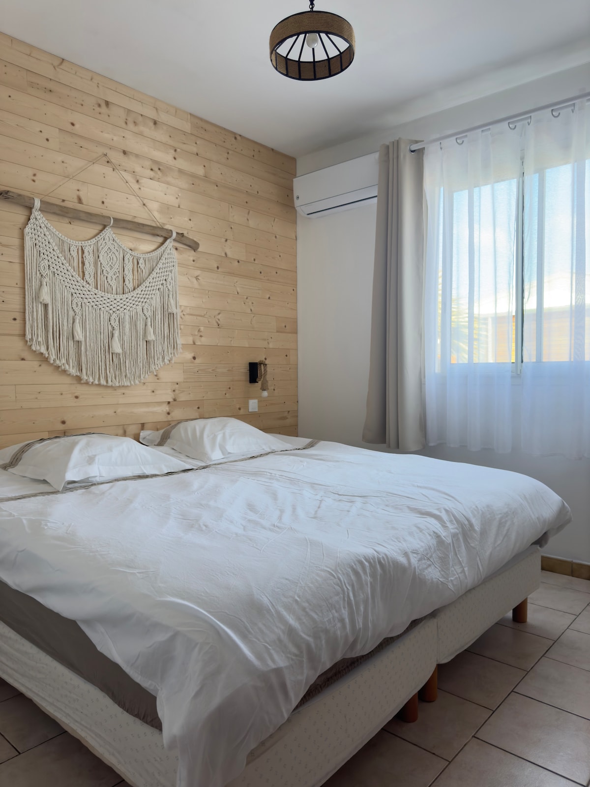 A bright and airy bedroom features a spacious bed with white linens. Natural light filters through a sheer curtain-covered window. Wooden paneling and a decorative textile piece enhance the room's warmth, while an air conditioning unit is positioned on the wall for comfort.