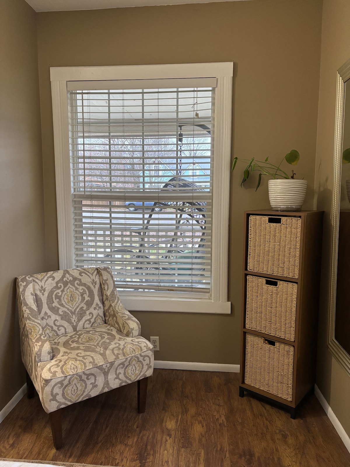 A corner nook features a patterned chair positioned beside a window with adjustable blinds. A multi-drawer storage unit stands next to the chair, showcasing a neutral design. A potted plant sits atop the storage unit, adding a touch of greenery.