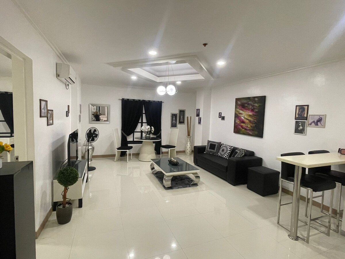 An open living area is showcased with modern furnishings and glossy tile floors. A black sectional sofa faces a coffee table, while a dining table with stools is positioned nearby. Walls are adorned with framed artwork, and dark curtains frame the windows.
