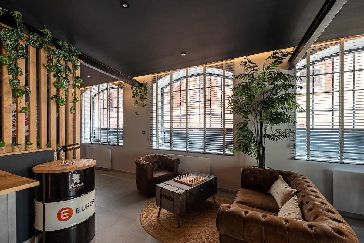 A modern living area is showcased with large windows offering ample natural light. Two brown leather sofas flank a rustic coffee table, while a chessboard adds a playful touch. Greenery is incorporated with potted plants, contributing to a relaxed ambiance.
