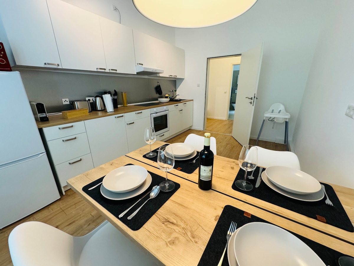 The kitchen area showcases a modern dining table set for four, featuring plates and wine glasses. Minimalist cabinetry and a spacious countertop are visible, along with kitchen appliances. Light wood flooring complements the overall design, creating an inviting and functional space.